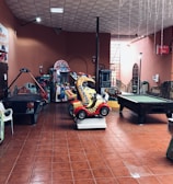 An indoor arcade area featuring various gaming machines, including an air hockey table and a ride-on toy car. The room has a tiled floor and several colorful gaming devices scattered around. Chairs and arcade signage are visible, creating a playful and entertainment-focused atmosphere.