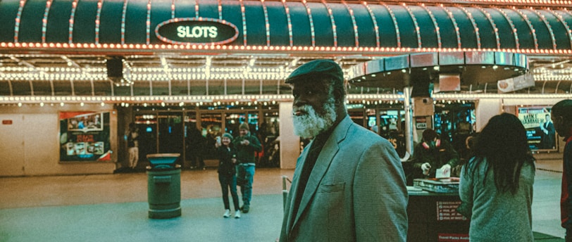 An older man with a gray beard and wearing a suit and hat stands in front of a brightly lit casino. The background features a large illuminated sign reading 'CASINO' and 'SLOTS' among other words, with numerous lights outlining the entrance. Several other people are visible in the background, engaged in various activities, some walking, some standing, and one looking at something on display.