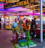 A vibrant amusement arcade featuring various games and bright neon signs. Mechanical horse rides are in the foreground, surrounded by claw machines with plush toys. The entrance is adorned with signs for a cinema and arcade, creating a lively and colorful atmosphere.