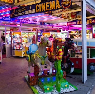 A vibrant amusement arcade featuring various games and bright neon signs. Mechanical horse rides are in the foreground, surrounded by claw machines with plush toys. The entrance is adorned with signs for a cinema and arcade, creating a lively and colorful atmosphere.
