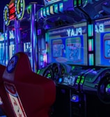 A vibrant arcade environment with several illuminated gaming machines. The image features arcade racing games with visible steering wheels, colorful control panels, and screens displaying game instructions. Bright LED lights in various colors illuminate the machines and surrounding area.