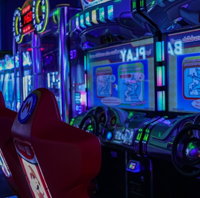 A vibrant arcade environment with several illuminated gaming machines. The image features arcade racing games with visible steering wheels, colorful control panels, and screens displaying game instructions. Bright LED lights in various colors illuminate the machines and surrounding area.